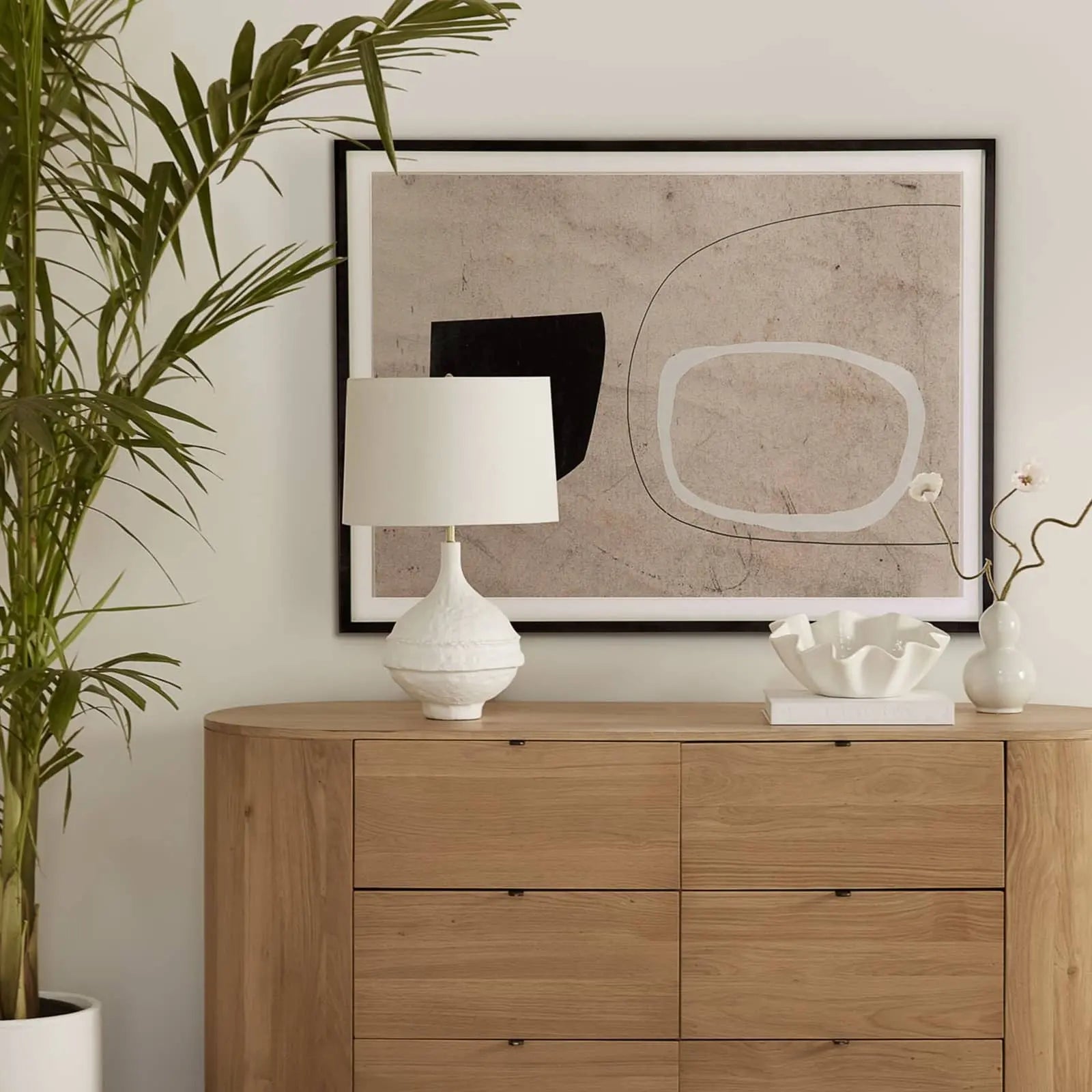 Wooden dresser with a lamp, abstract art, and decorative items in a room setting.