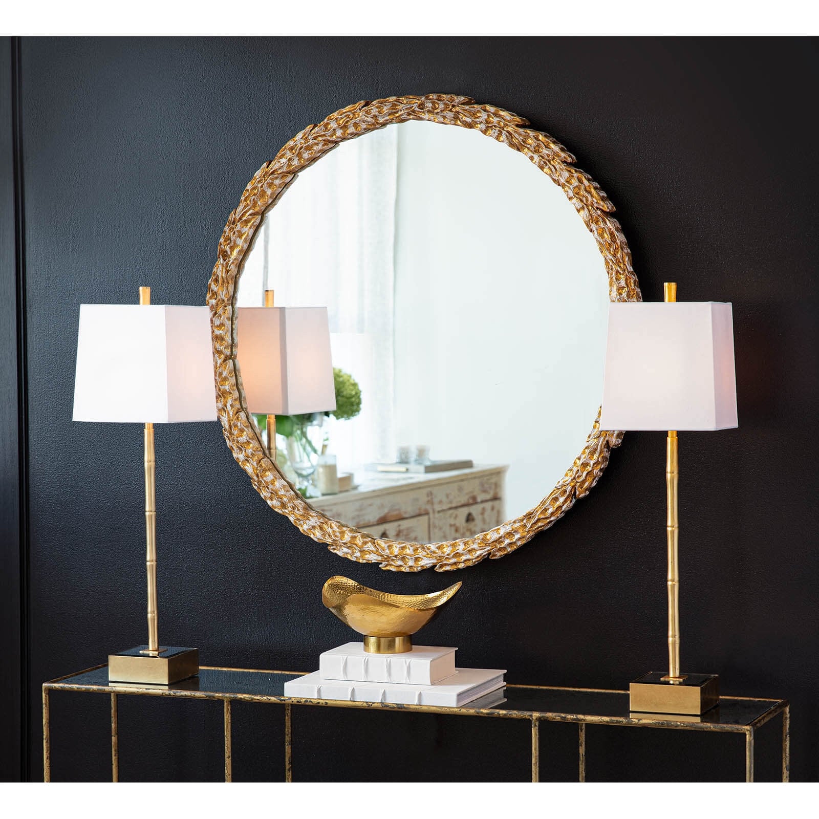 Round decorative mirror with gold frame on a dark wall with two table lamps.
