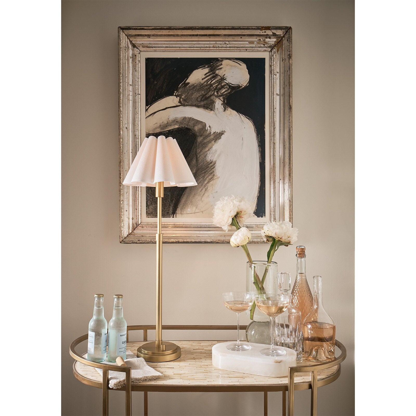 Decorative table with lamp, bottles, and glasses against a wall with framed art.