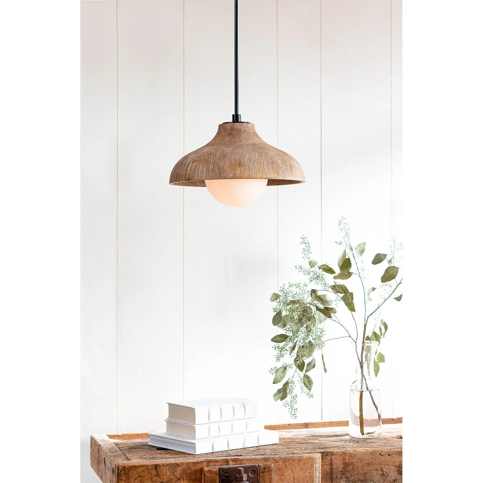 Wooden pendant light hanging above a wooden table with books and a plant against a white wall.