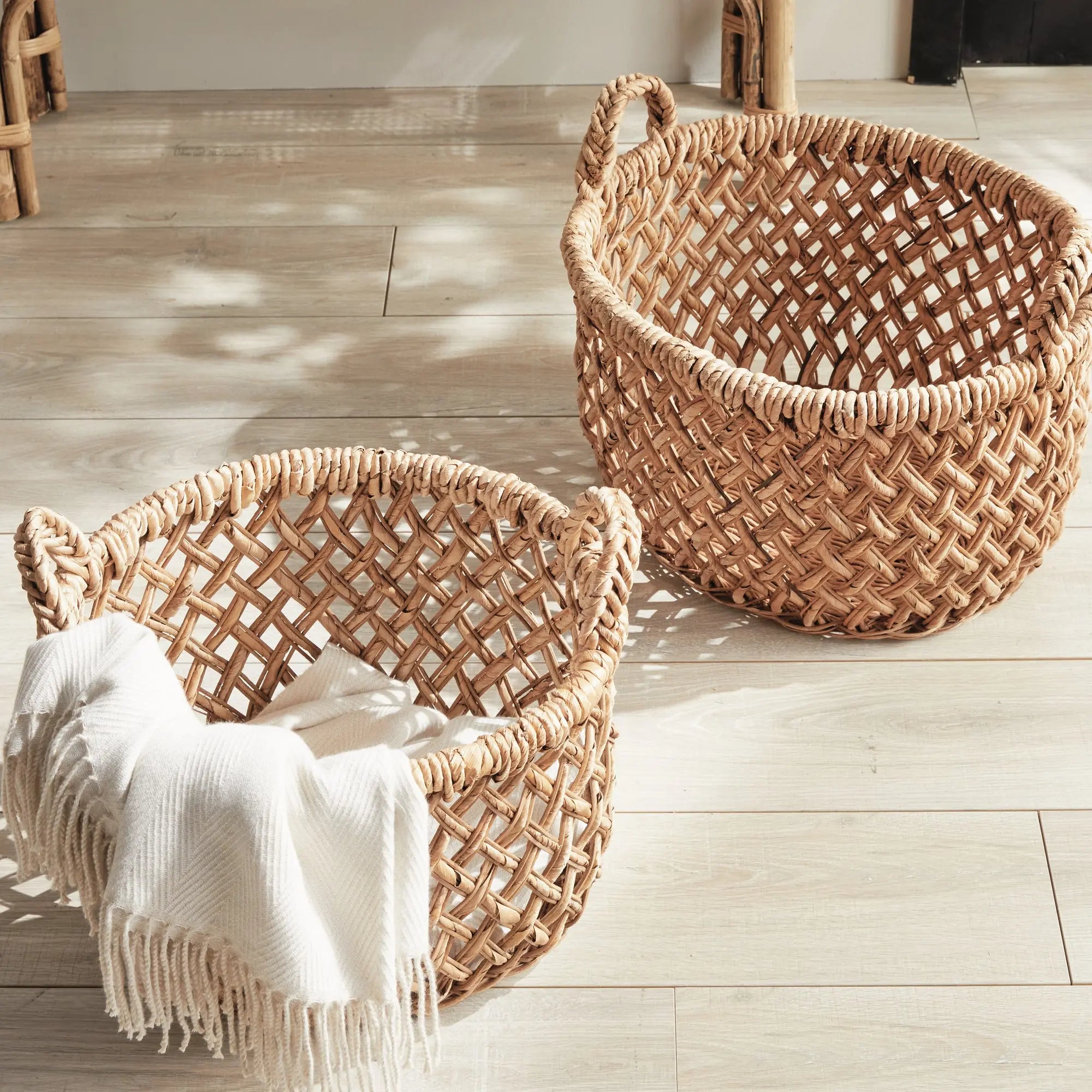 Emlyn Woven Storage Baskets with Handles – Set of 2 (Banana Leaf) - Two woven baskets on a wooden floor with a white blanket inside one of them.