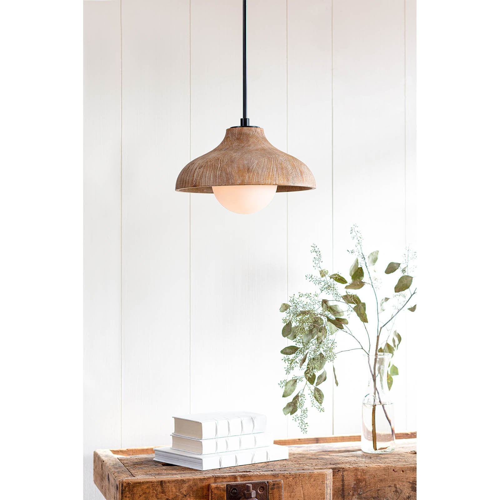 Wooden pendant light hanging above a wooden table with books and a plant against a white wall.