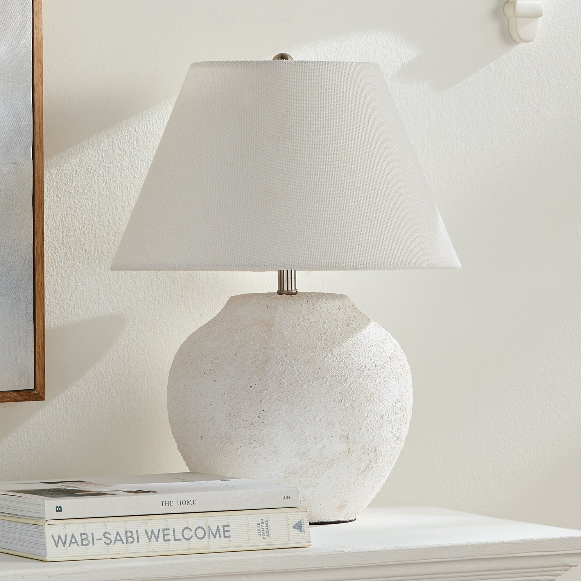Theo Lamp - White textured table lamp on a shelf with books underneath, against a light wall.